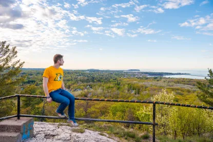 A man in a yellow T-shirt and jeans sits on a metal railing atop a rocky hill, looking out over a scenic view of a green forest, a town below, and a distant body of water under a partly cloudy sky.