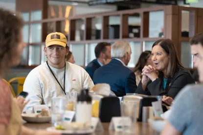 Student in yellow hat eating with board of trustee member in dining hall