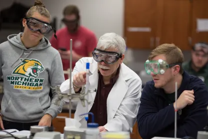 Students and professor interact in a chemistry lab wearing proper safety gear