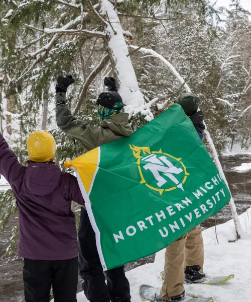 Students stand in winter gear, backs facing the camera, on the edge of a river, holding a Northern Michigan University flag behind their backs