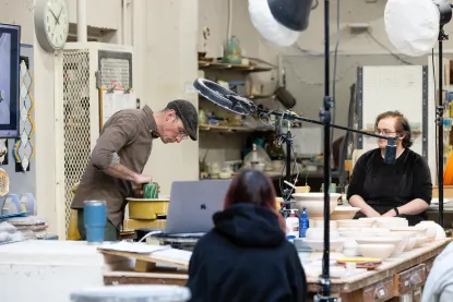 Three people are in a ceramics studio; one shapes clay on a pottery wheel, another watches while seated, and a third sits with their back to the camera. Pottery and equipment can be seen around the workspace.
