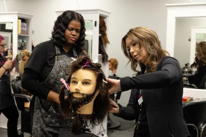 Two women in a salon practice hairstyling on a mannequin head with a beard. One woman styles the hair while the other observes, both focused on their work in a classroom-like salon setting.