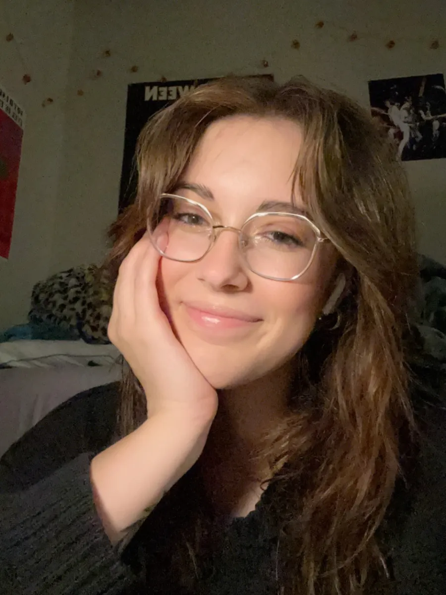 Student with brown hair and clear glasses, sitting in dorm room, smiling at the camera. There are decorations and posters on the wall behind her.