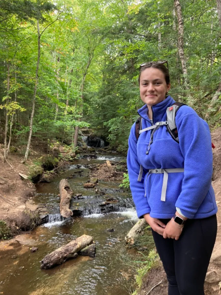 Student wearing blue jacket and backpack standing on the bank of a stream in front of a waterfall. 