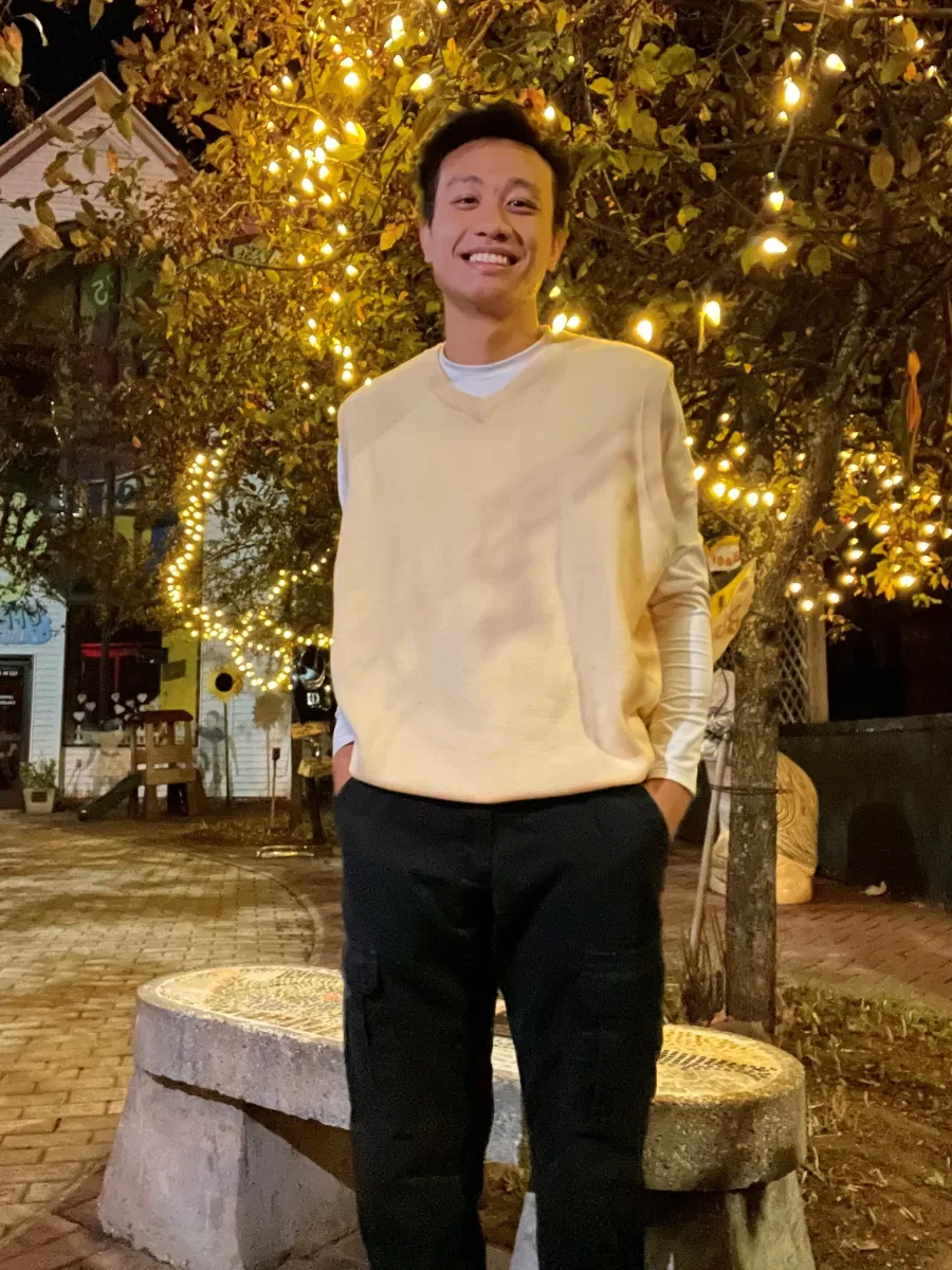 Student in white sweater/ black pants standing in front of a tree with yellow lights. 