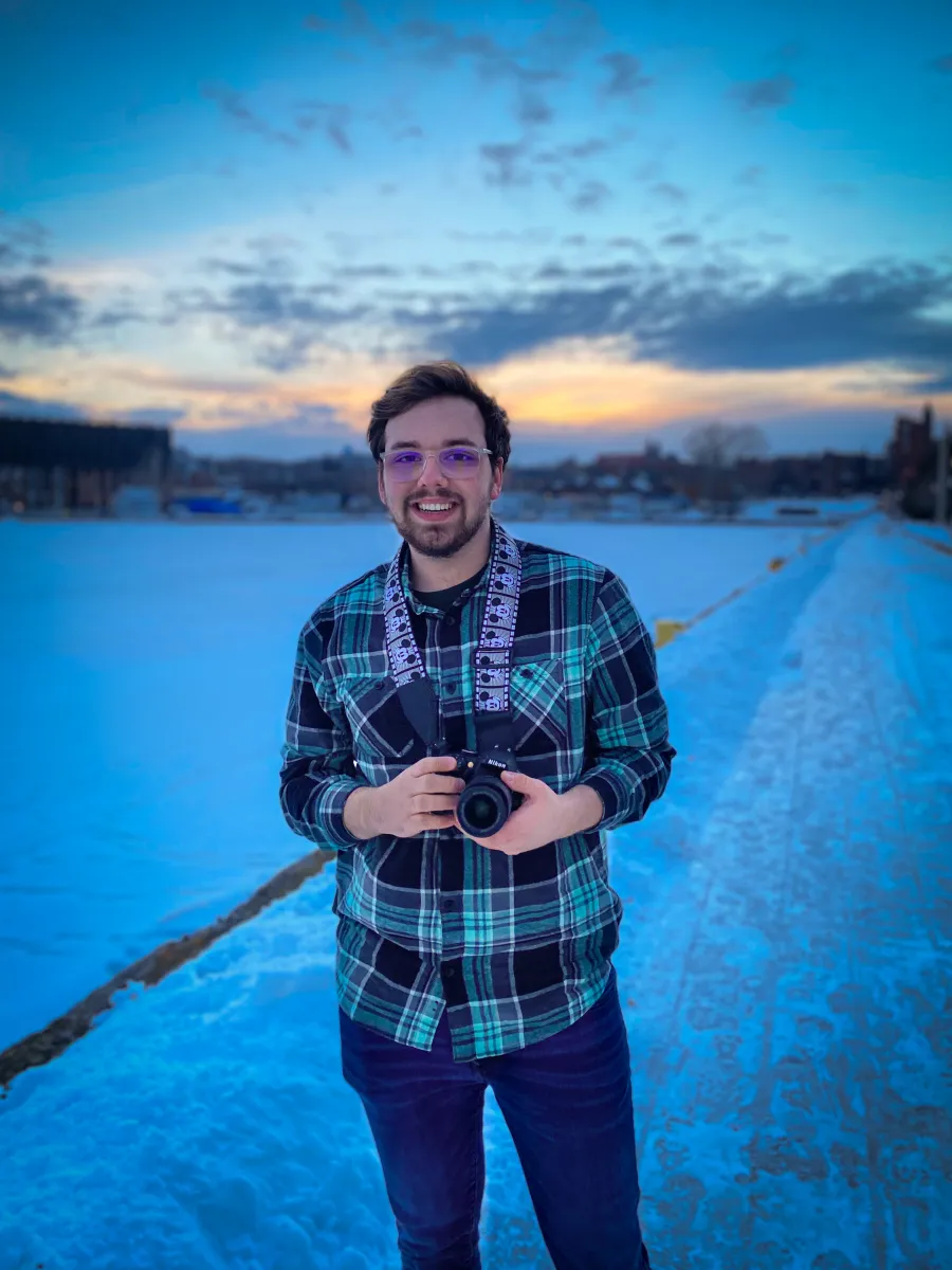 Student wearing green plaid, with a camera around his neck, standing next to the frozen lower harbor in downtown Marquette.