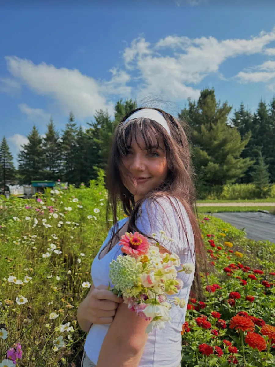 A student stands in a field of flowers wearing a white shirt with a white headband in brunette hair. The student holds a bouquet of flowers.  