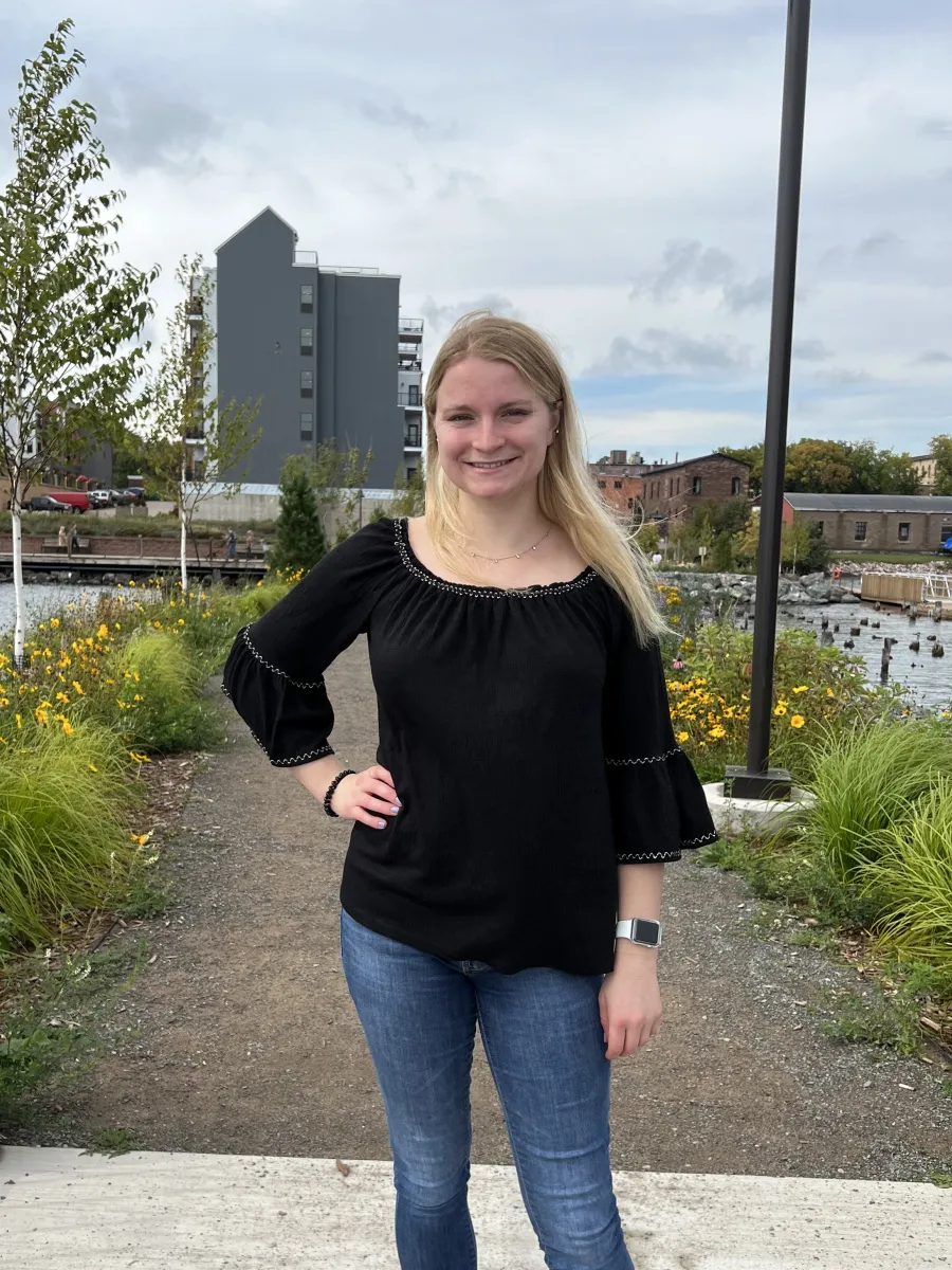 A blond student wearing a black blouse and blue jeans stands in front of a natural landscape facing the camera. 