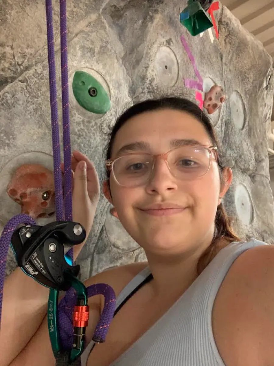 A student with brown hair and glasses smiles towards the camera. The angle is a selfie showing off the student climbing the rock wall. 