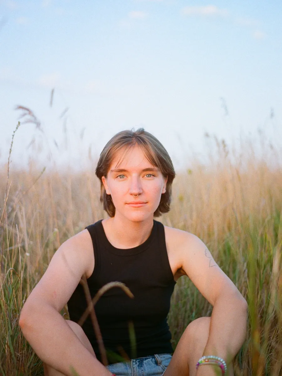 A student with short dirty blond hair sits in a field of tall grasses facing the camera. They are earing a black tank top and blue shorts. 