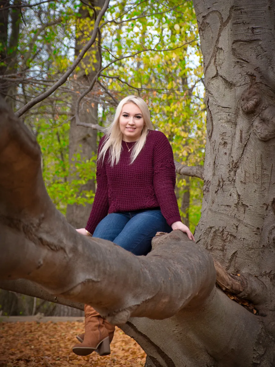 A student with blonde hair sits in a tree facing the camera. They are wearing a burgundy sweater and blue jeans. 
