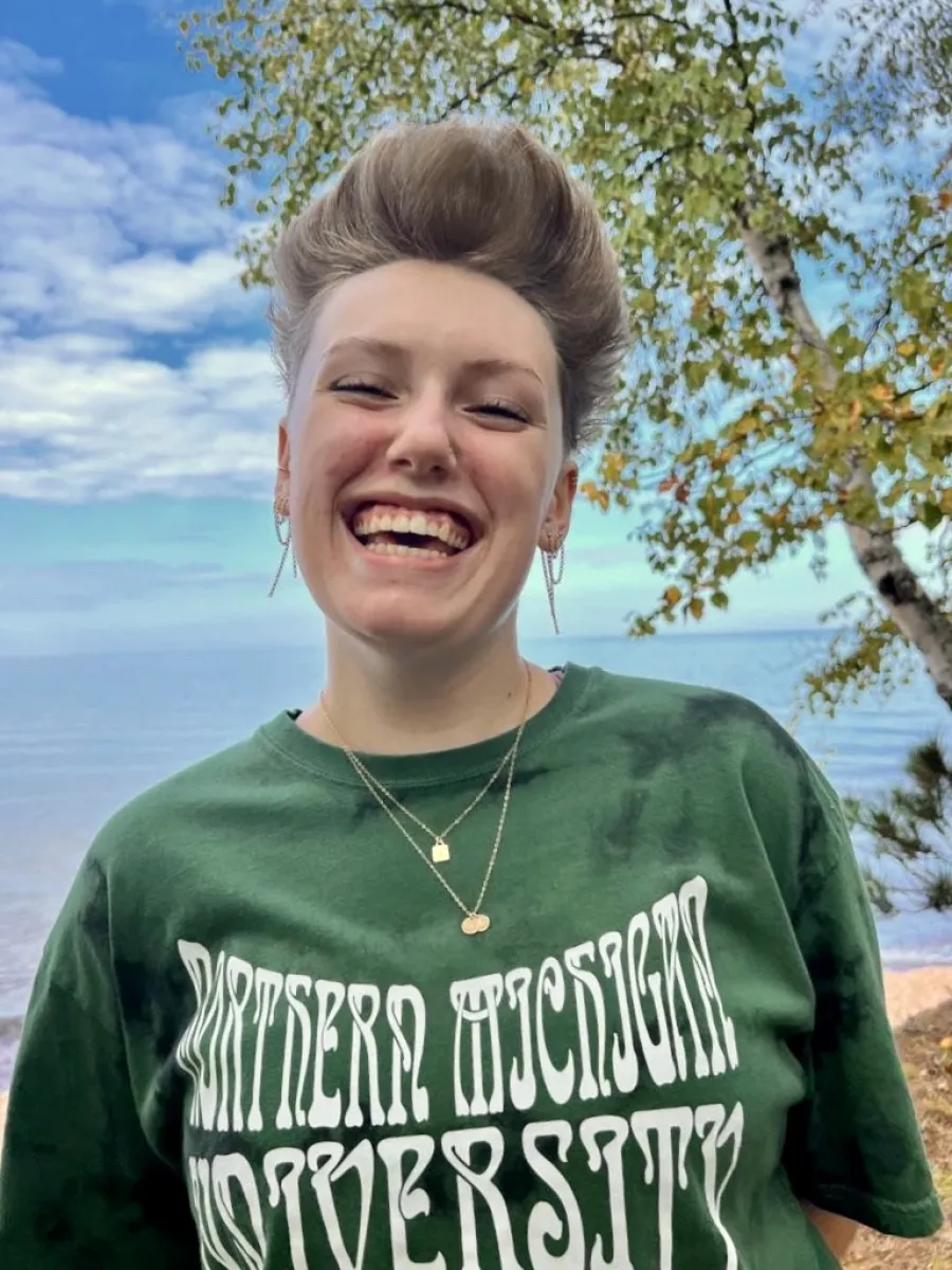A student with short hair faces the camera and smiles. Lake Superior is in the background and they are wearing a green and white Northern Michigan University shirt. 