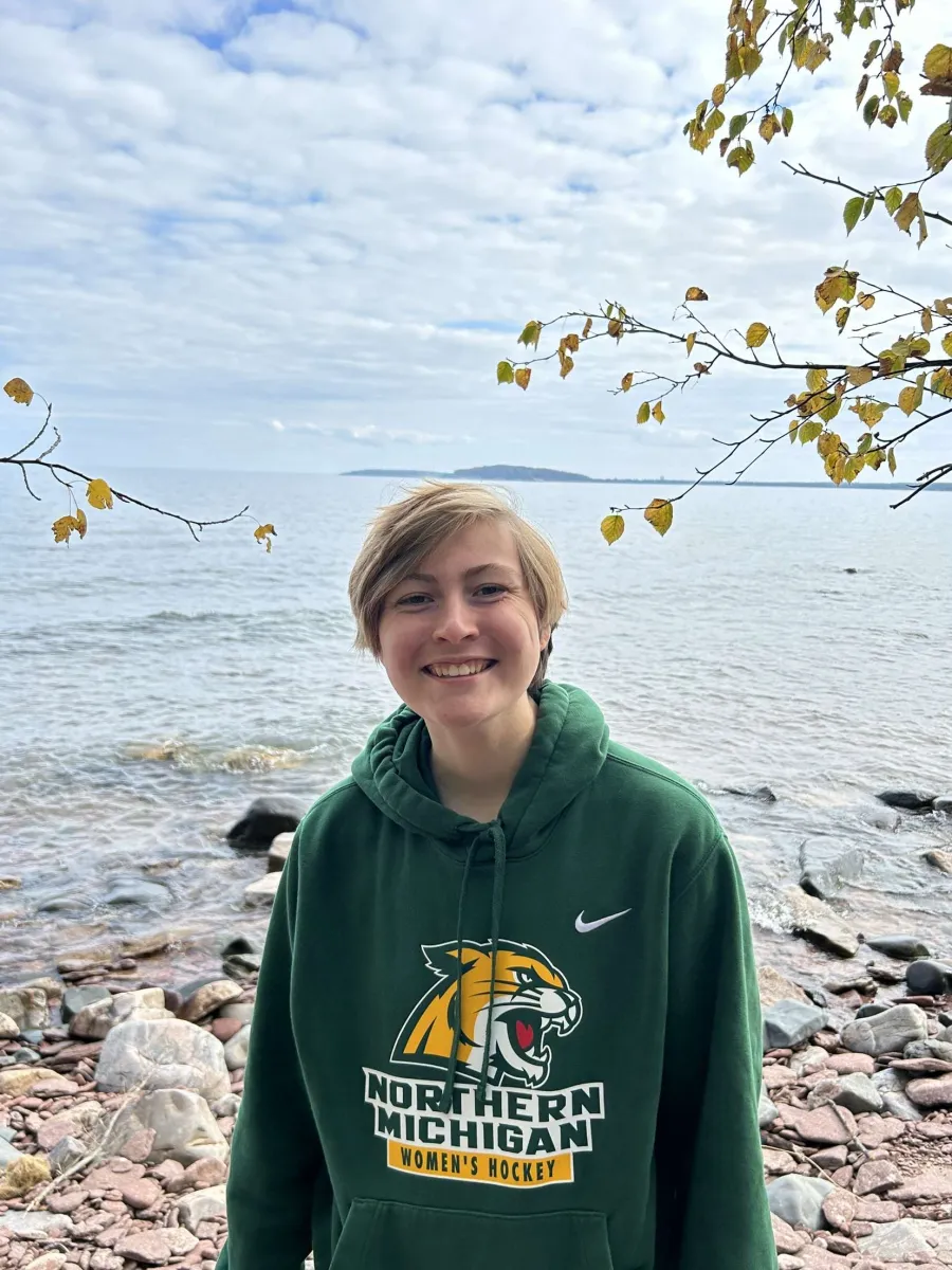 A student with short blond hair stands facing the camera with a smile. They are wearing a green NMU Wildcat hooded sweatshirt and Lake Superior is in the background. 