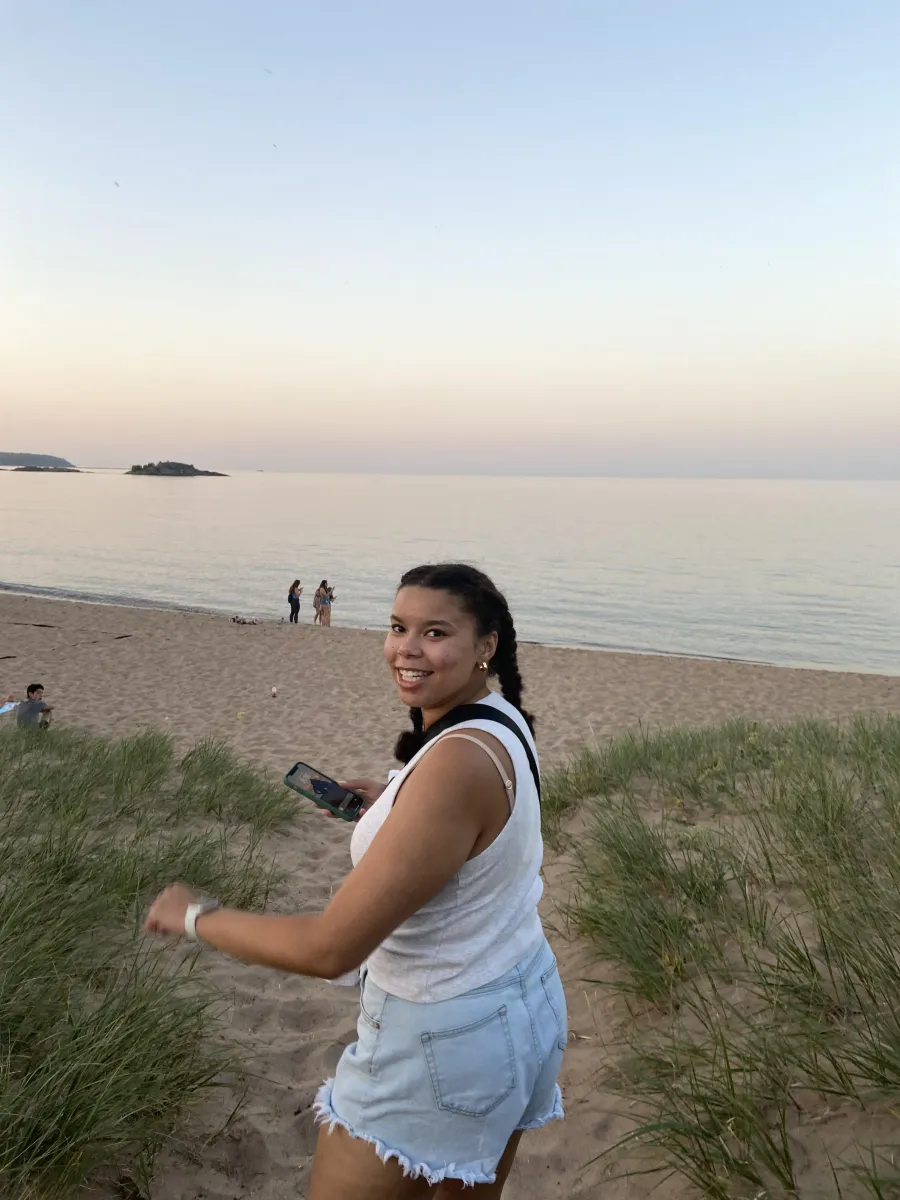 A student is featured walking on a beach towards the Lake Superior Shoreline. She is turning back slightly to face the camera with a smile. 