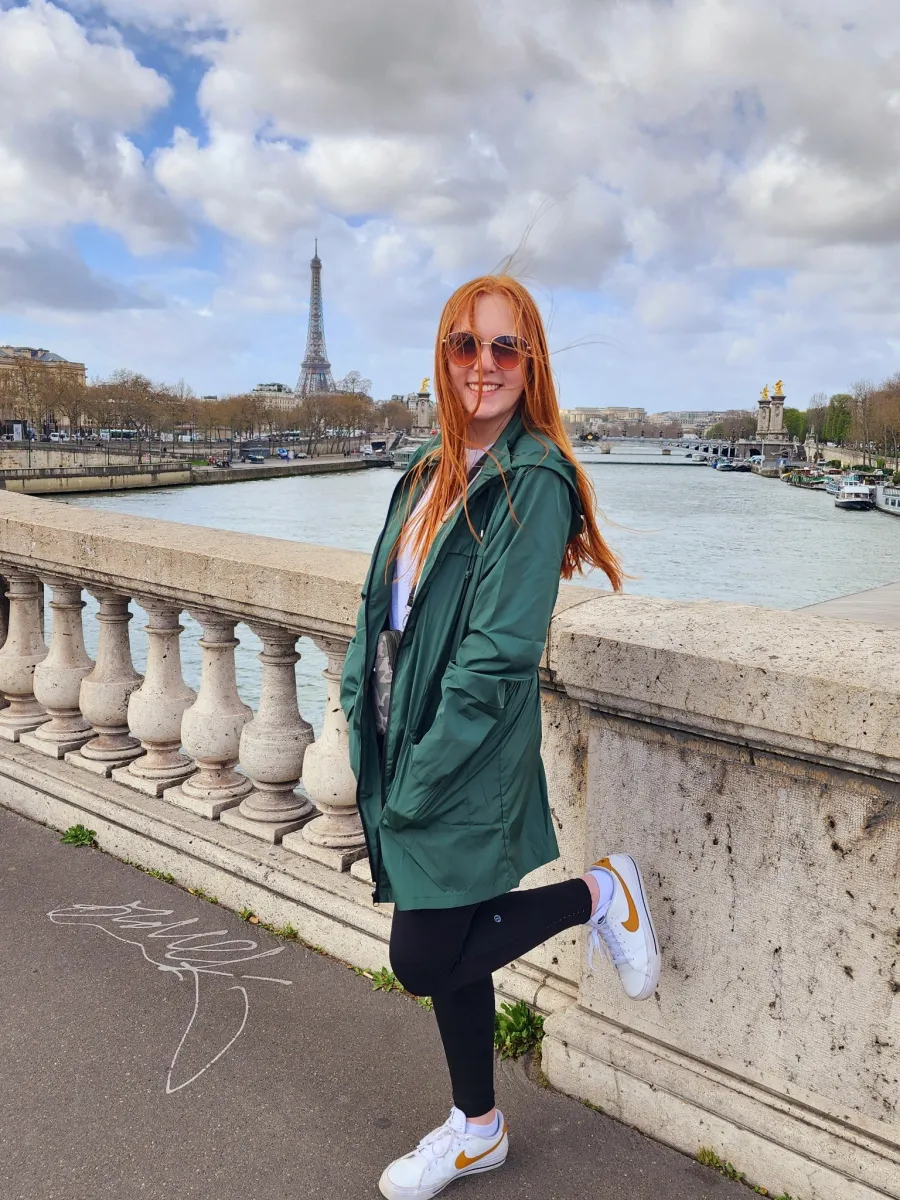 A student with red hair stands in front of a river, wearing a green jacket and black pants. The Eiffel tower is in the background. 