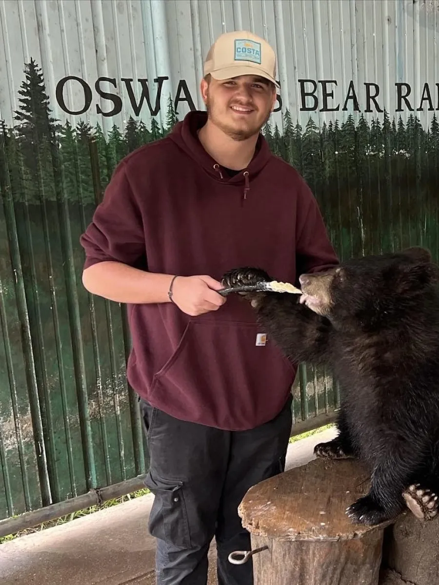 A student wearing a dark red sweatshirt and tan hat faces the camera with a smile. He is feeding a chubby black bear cub a treat. The background is a mural of trees that reads "Oswald's Bear Ranch"