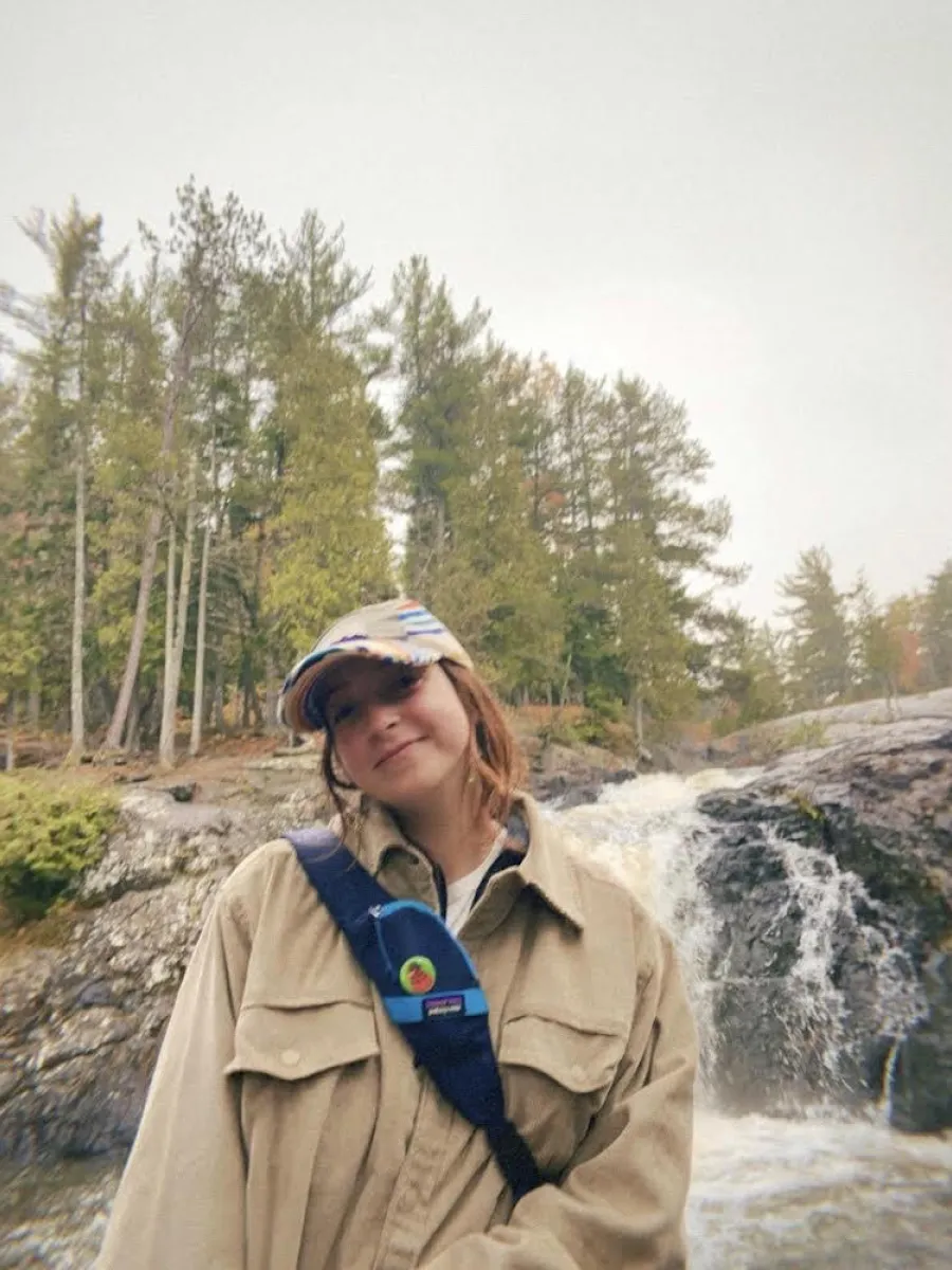 A student wearing a tan jacket and hat, with a blue sling bag hanging off her shoulder stands in front of a waterfall. 