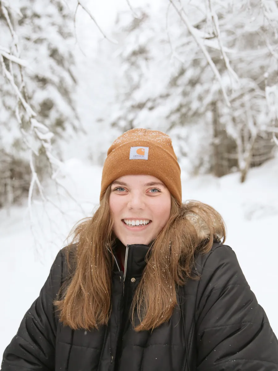 A student with long brown hair, wearing a tan beanie hat and black jacket stands outdoors with a snowy background. 