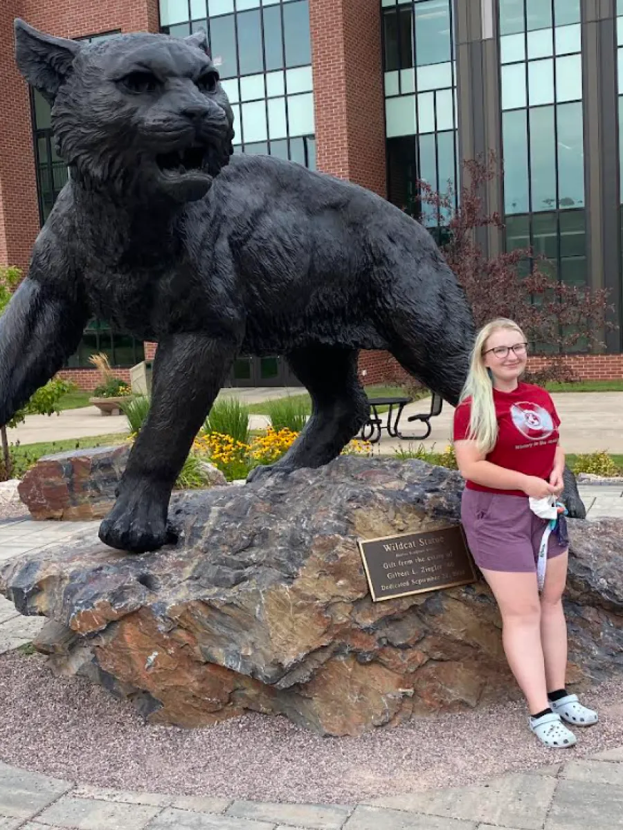 A student with blond hair and red shirt is leaning up against the rock that holds a Wildcat Statue.