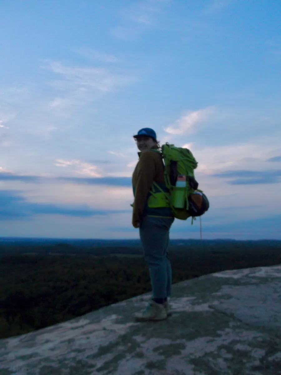 Delaney in a hat and hiking gear, standing on top of Hog Back Mountain with a backpack