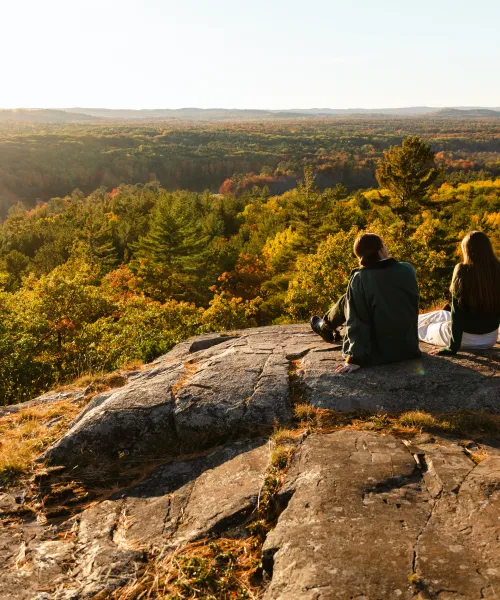 Three students sit on a rocky outcropping staring off into a sunset over a forest