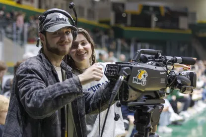 Two broadcasting students smile at the camera while live broadcasting an NMU basketball game