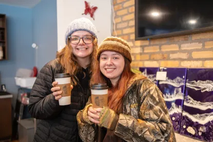 Two women in winter hats hold to go coffee cups and smile at the camera