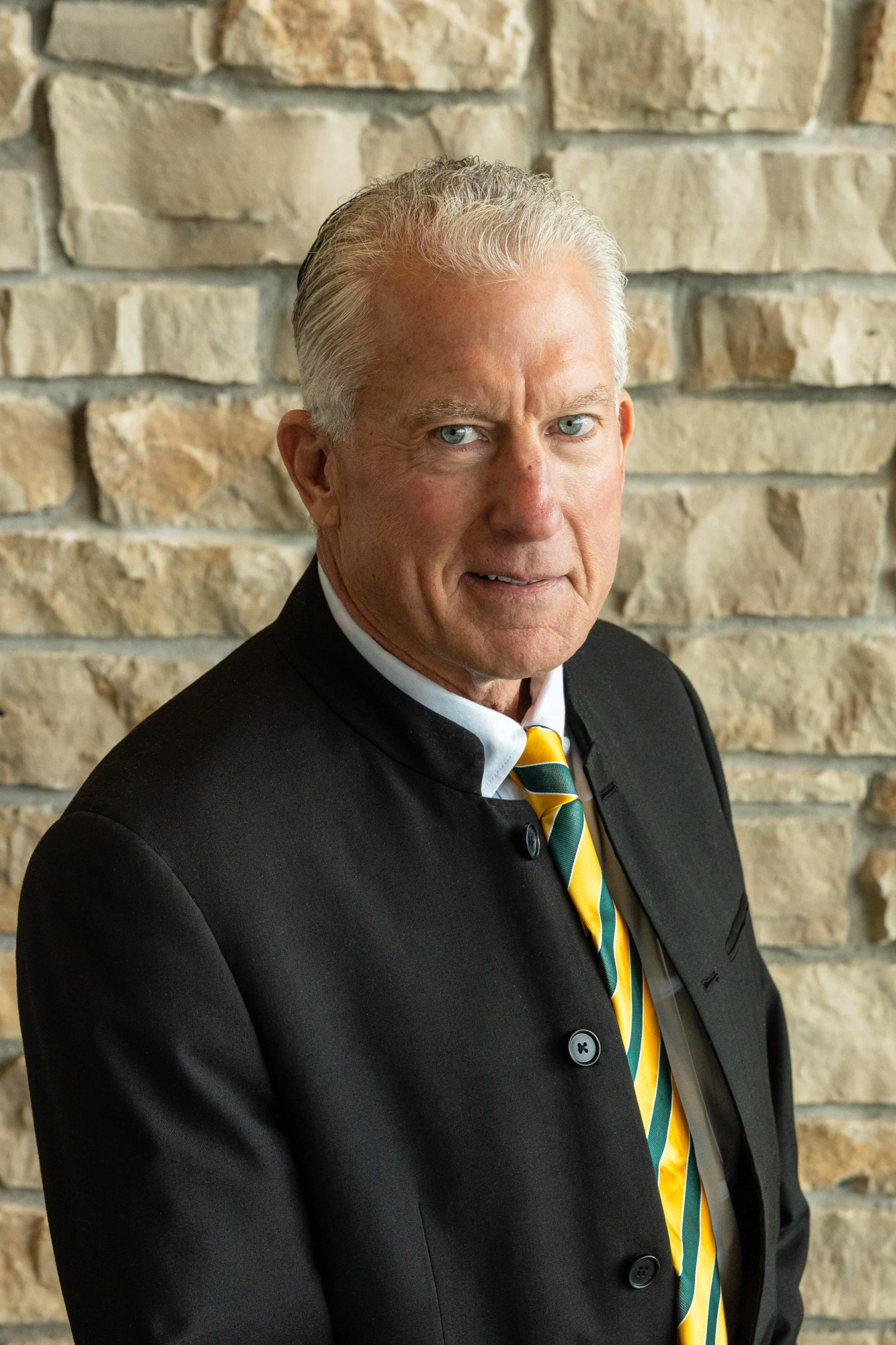 Bill Sickert smiles against a stone backdrop wearing a green and gold tie and a black sportcoat