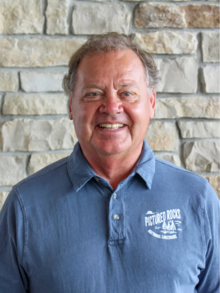 A headshot photo of John Madigan smiling against a stone backdrop with natural lighting in a polo tee shirt.