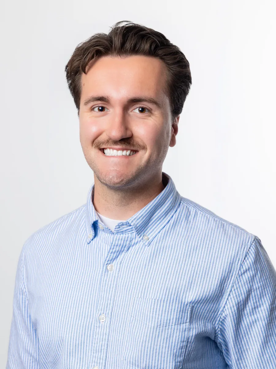 A man in a light blue button up smiles against a white background.