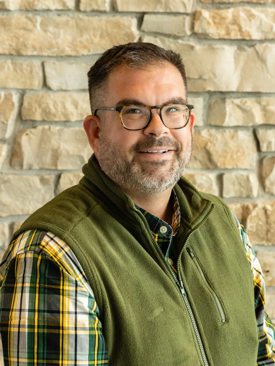 A headshot photo of a man with short hair and a beard and glasses wearing a fleece green vest and a flannel shirt smiling against a stone backdrop with natural lighting in a polo tee shirt.