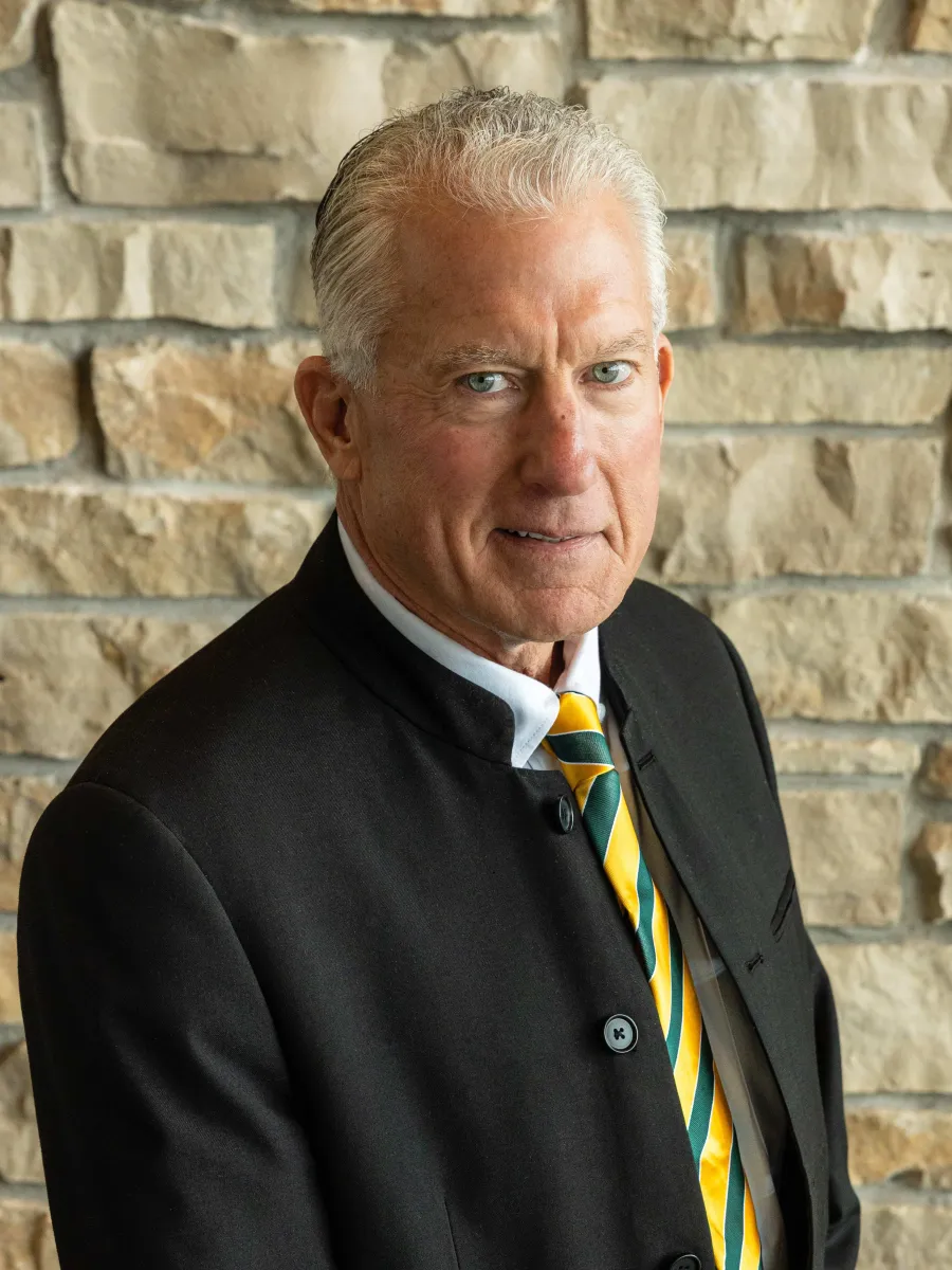 A headshot photo of Bill Sickert in a sport coat, a white button down shirt, and a green and yellow tie, smiling against a stone backdrop with natural lighting.