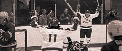 An NMU Wildcats player raises his arms to celebrate a goal as teammates and fans behind the glass cheer in jubilation.