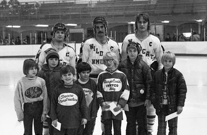 Young children in youth hockey jerseys pose on ice with three NMU Wildcats hockey players in a vintage black-and-white arena photo.
