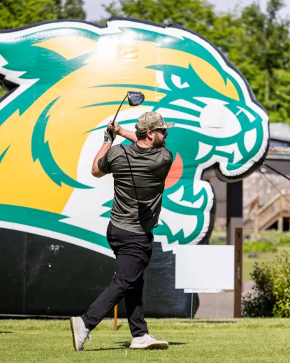 A golfer follows through on a drive in front of a large NMU Wildcats inflatable logo at an outdoor golf outing.