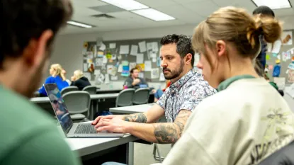 A man in a classroom clicks on a student's laptop surveying their work at the student's desk.