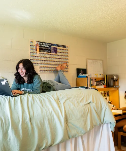 A person laying on their stomach on a bed in a dorm room with light green bedding, a laptop open in front of them. Another sitting at a desk.