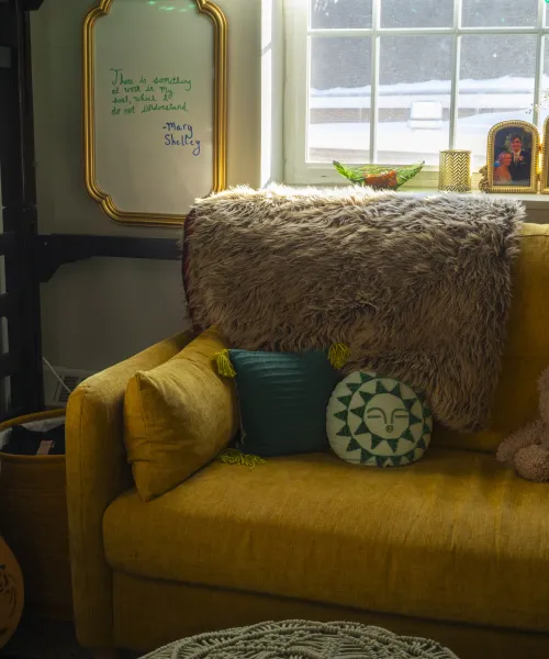 A yellow sofa with green throw pillows and a teddy bear in front of a window