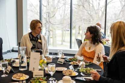 Three women sit at a round table set for a meal, engaged in conversation. 