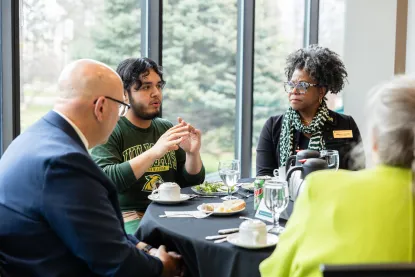 Four people sit around a table with coffee cups and plates, engaged in conversation by large windows with trees outside.