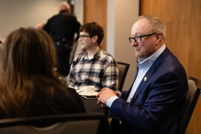 A man in a blue blazer and glasses sits at a table with two others.
