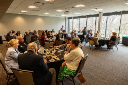 A group of people sit at round tables in a conference room with large windows, eating, talking, and listening to a speaker at a podium in a formal.