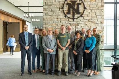 A group of twelve professionally dressed people stand smiling in front of a stone wall with a large "N" logo at a conference venue.