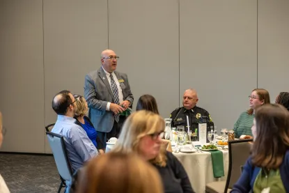 	 A man in a gray suit stands and speaks to a group seated around tables at an event