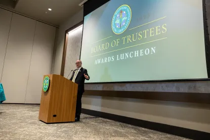 A man in a suit speaks at a podium during a Board of Trustees Awards Luncheon, with a large presentation screen displaying the event's title and official seal behind him.