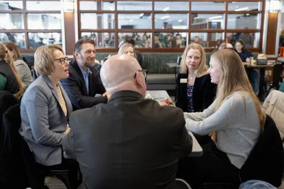A group of five adults sits around a table engaged in conversation in a busy indoor setting with more people and glass windows in the background.