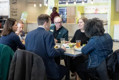 Six people sit around a table in a modern cafeteria, eating lunch and having a conversation.