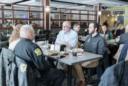 A group of people sit around a table in a bright cafeteria, eating and talking together. 