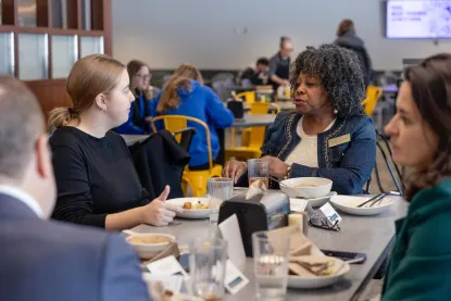 A group of people sit around a table in a bright cafeteria, eating and talking together. 