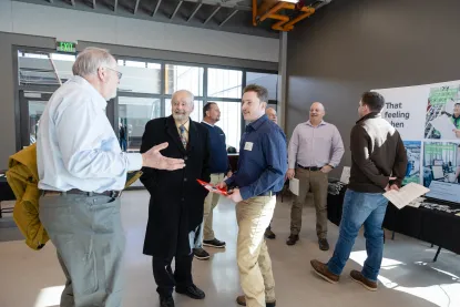 A group of men talk and mingle at an indoor event; some stand near a table with brochures while others converse in the foreground.